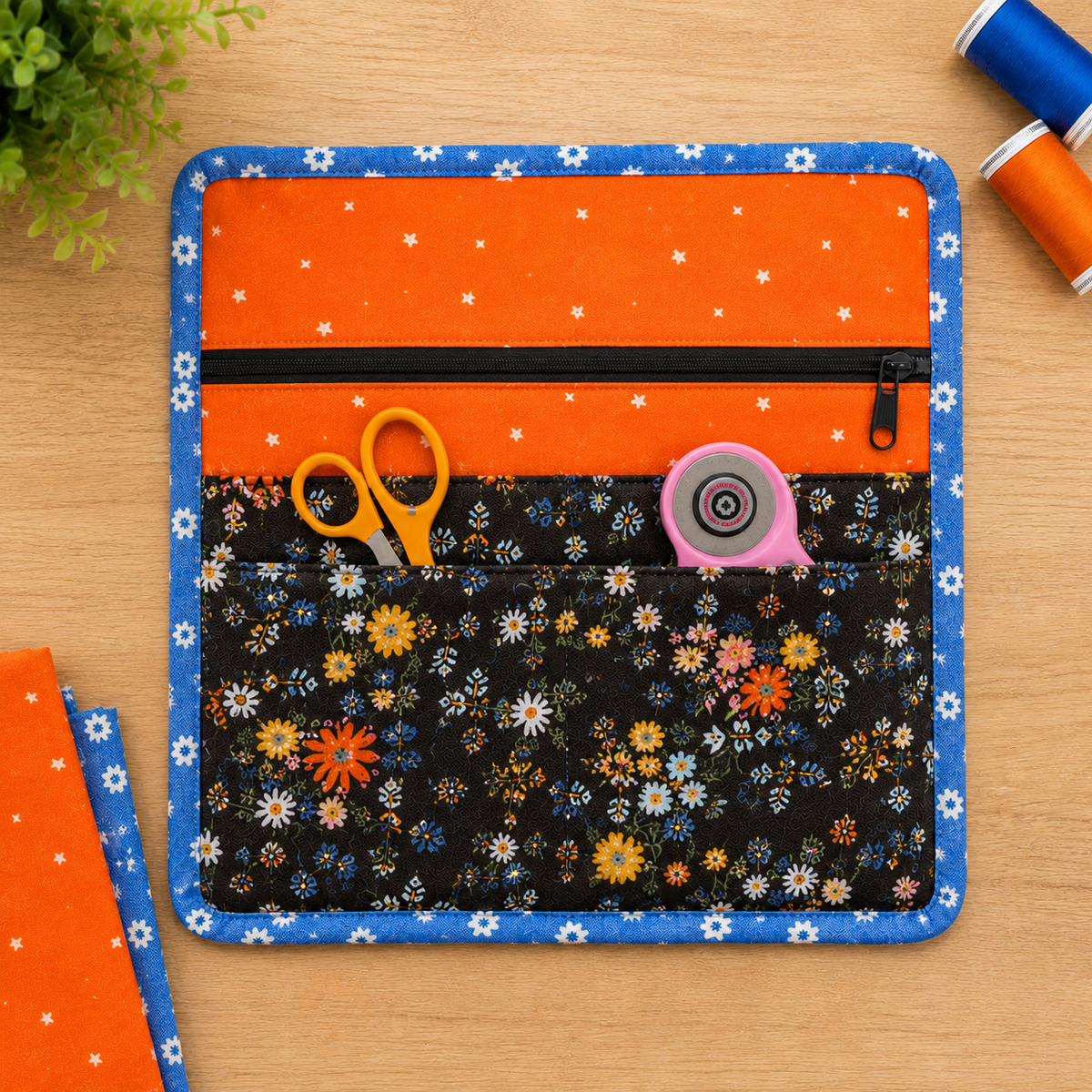 An orange and blue fabric organizer pouch with pockets, shown on a wooden tabletop with sewing supplies like scissors, a rotary cutter, and spools of thread.