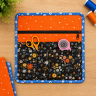 An orange and blue fabric organizer pouch with pockets, shown on a wooden tabletop with sewing supplies like scissors, a rotary cutter, and spools of thread.