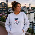 Woman wearing a gray sweatshirt with a graphic design, standing in front of a lighthouse and 'Welcome to Rhode Island' sign.