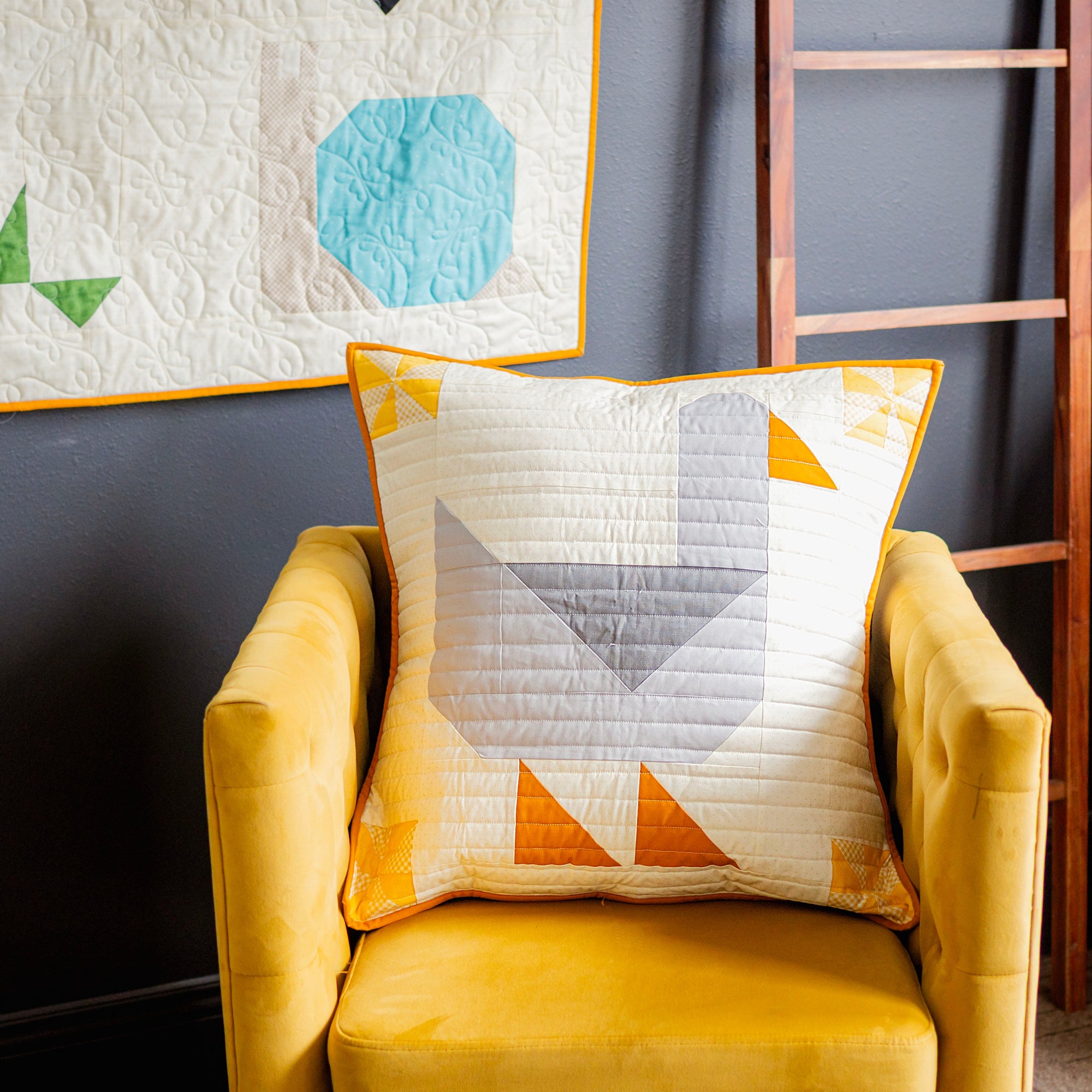 Yellow armchair with a geometric-patterned pillow against a dark wall with a quilt hanging above.