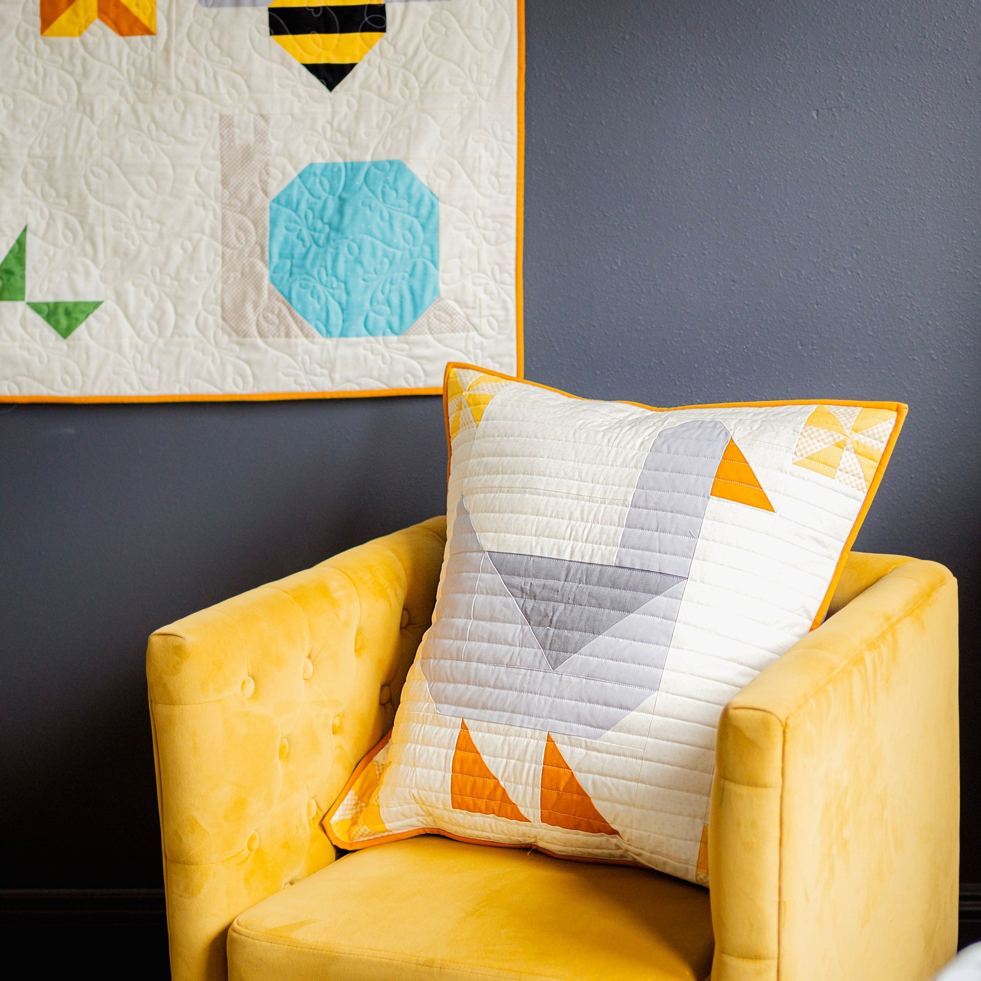 Yellow armchair with a geometric-patterned goose pillow against a dark wall with a quilt hanging above.