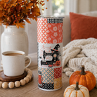 A tall insulated tumbler with a clear lid and straw stands on a wooden table in a cozy autumn setting. The tumbler features a patchwork design with orange floral, geometric, and Halloween-themed prints, including a sewing machine, witch hat, spools, and jack-o'-lanterns.