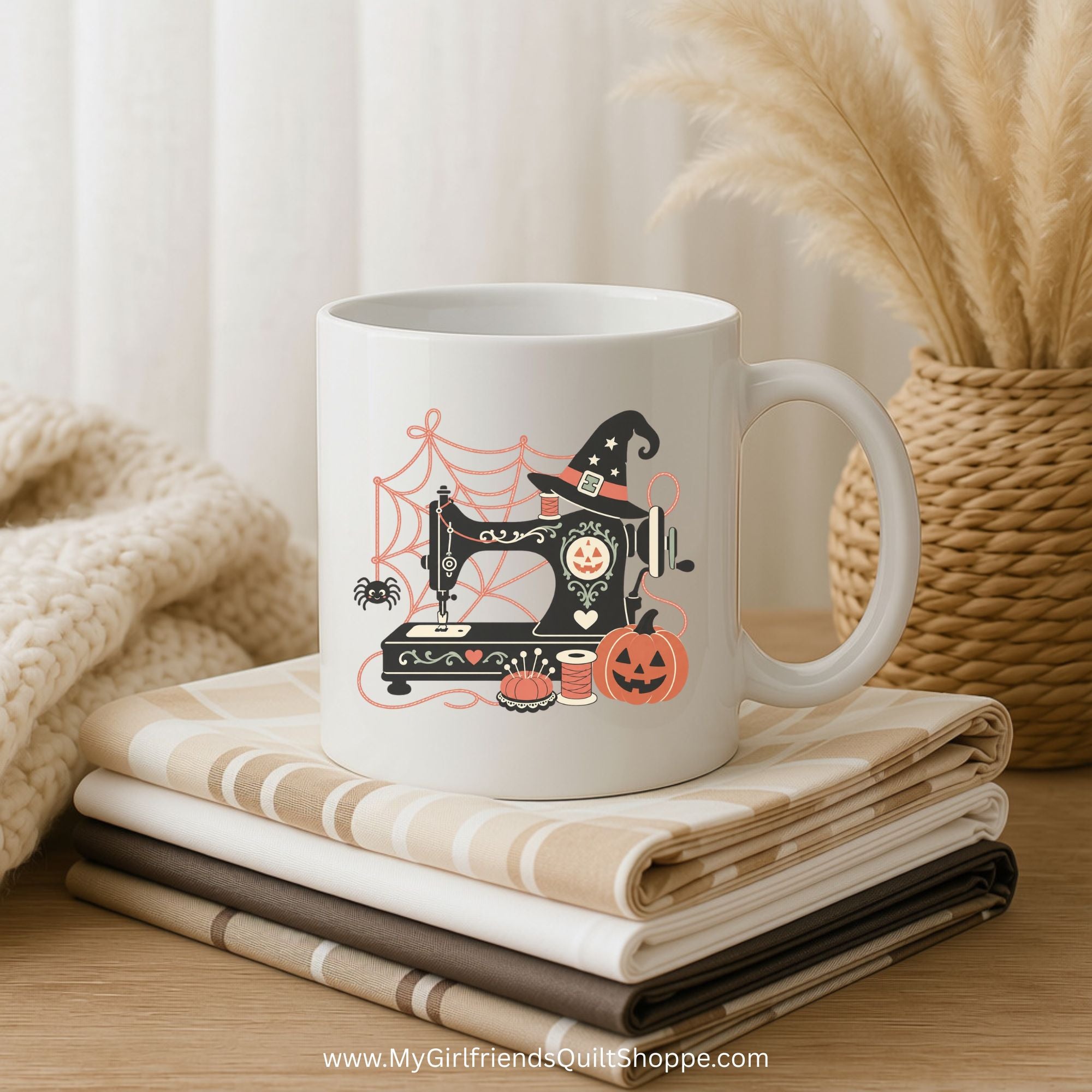 A white ceramic mug with a Halloween-themed sewing machine illustration sits on a stack of folded beige and brown quilts on a wooden table. A knitted blanket is on the left, and a wicker basket with dried pampas grass is on the right.