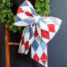 Quilted blanket with red, white, and blue pattern draped over a wooden ladder with a green wreath on a dark background