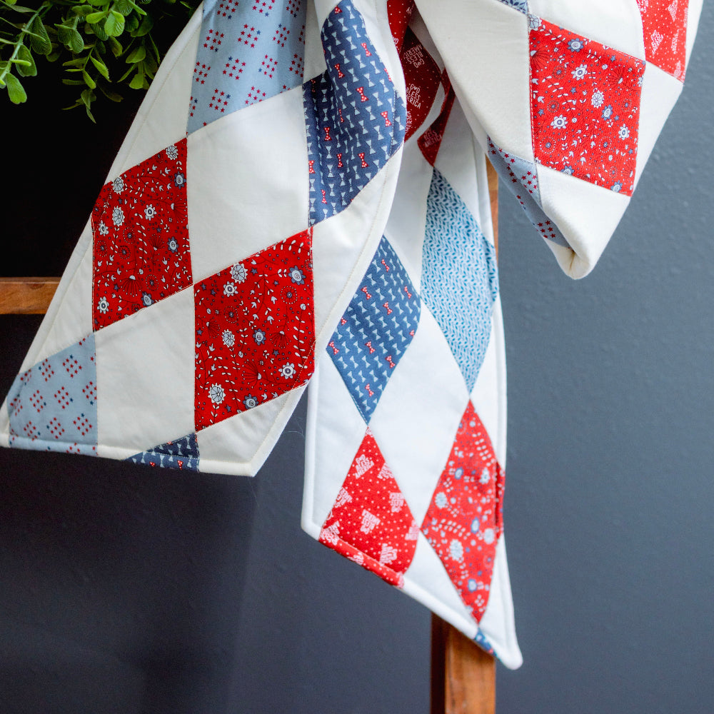 Quilt with red, white, and blue diamond pattern draped over a chair against a dark background.
