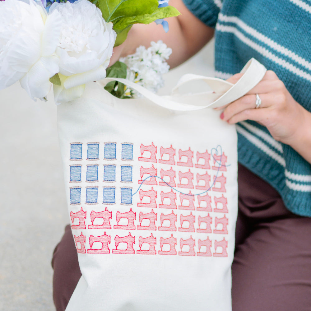 Person holding a tote bag with a pattern, surrounded by flowers and wearing a blue sweater.