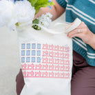 Person holding a tote bag with a pattern, surrounded by flowers and wearing a blue sweater.