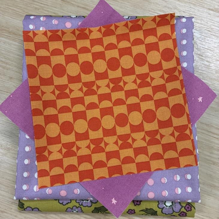 A stack of patterned fabric squares on a light wood surface. The top fabric has an orange and yellow geometric checkerboard pattern, layered over purple fabrics with white polka dots and a green floral patterned fabric underneath.