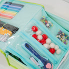 A mint green and lime green zippered craft organizer pouch filled with sewing and crafting supplies, including colorful pens, buttons, pom-poms, and a rotary cutter, shown on a white background.