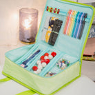 An open light blue and lime green fabric pen and craft organizer case, filled with pens, markers, beads, and small craft supplies, displayed on a tabletop with a blurred candle in the background.