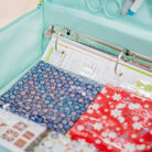 A mint green zippered organizer pouch holding a clear binder with patterned paper inserts, including blue and red floral sheets.