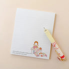 A lined notepad with a cute illustrated character and small quote at the bottom, paired with a pastel yellow pen that has a matching patterned sleeve, arranged on a warm beige background.