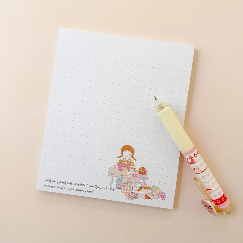 A lined notepad with a cute illustrated character and small quote at the bottom, paired with a pastel yellow pen that has a matching patterned sleeve, arranged on a warm beige background.