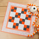 A handmade quilted baby blanket with a checkered pattern of orange, black, and white squares, styled on a light wood background next to a white pom-pom toy.