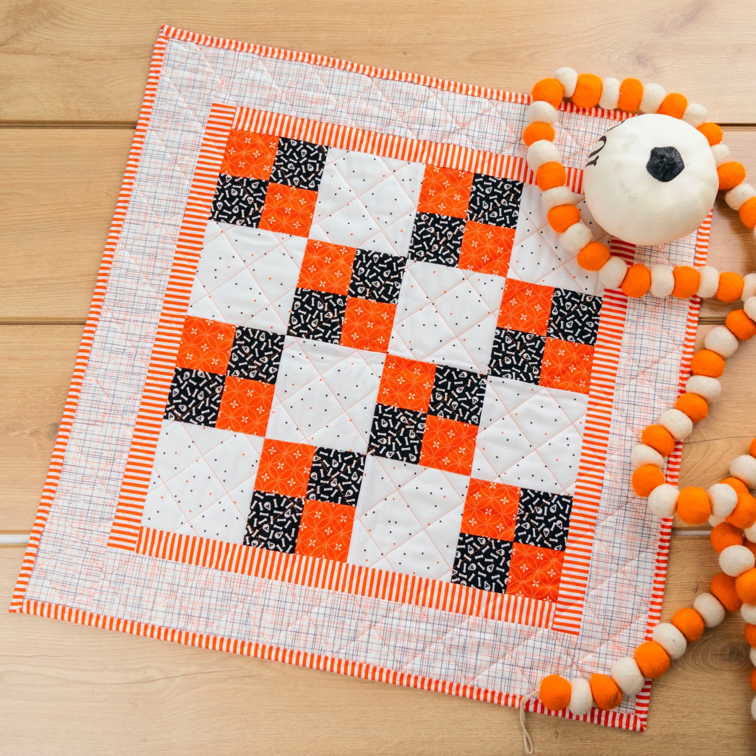 A handmade quilted baby blanket with a checkered pattern of orange, black, and white squares, styled on a light wood background next to a white pom-pom toy.