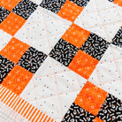 A close-up of a quilted fabric with a patchwork design featuring orange and black patterned squares on a white background, finished with an orange and white striped border.