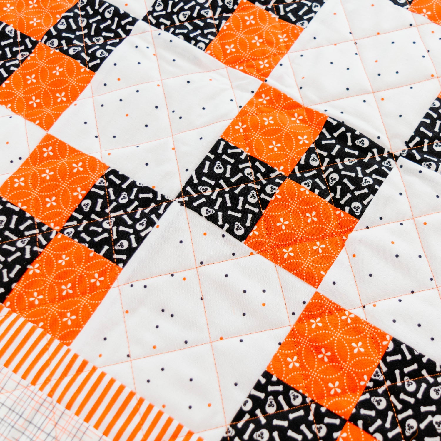 A close-up of a quilted fabric with a patchwork design featuring orange and black patterned squares on a white background, finished with an orange and white striped border.
