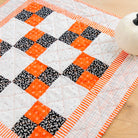 A handmade quilted table runner or placemat featuring a geometric patchwork design of orange, black, and white fabrics, styled on a light wood surface next to a small white decorative pumpkin.