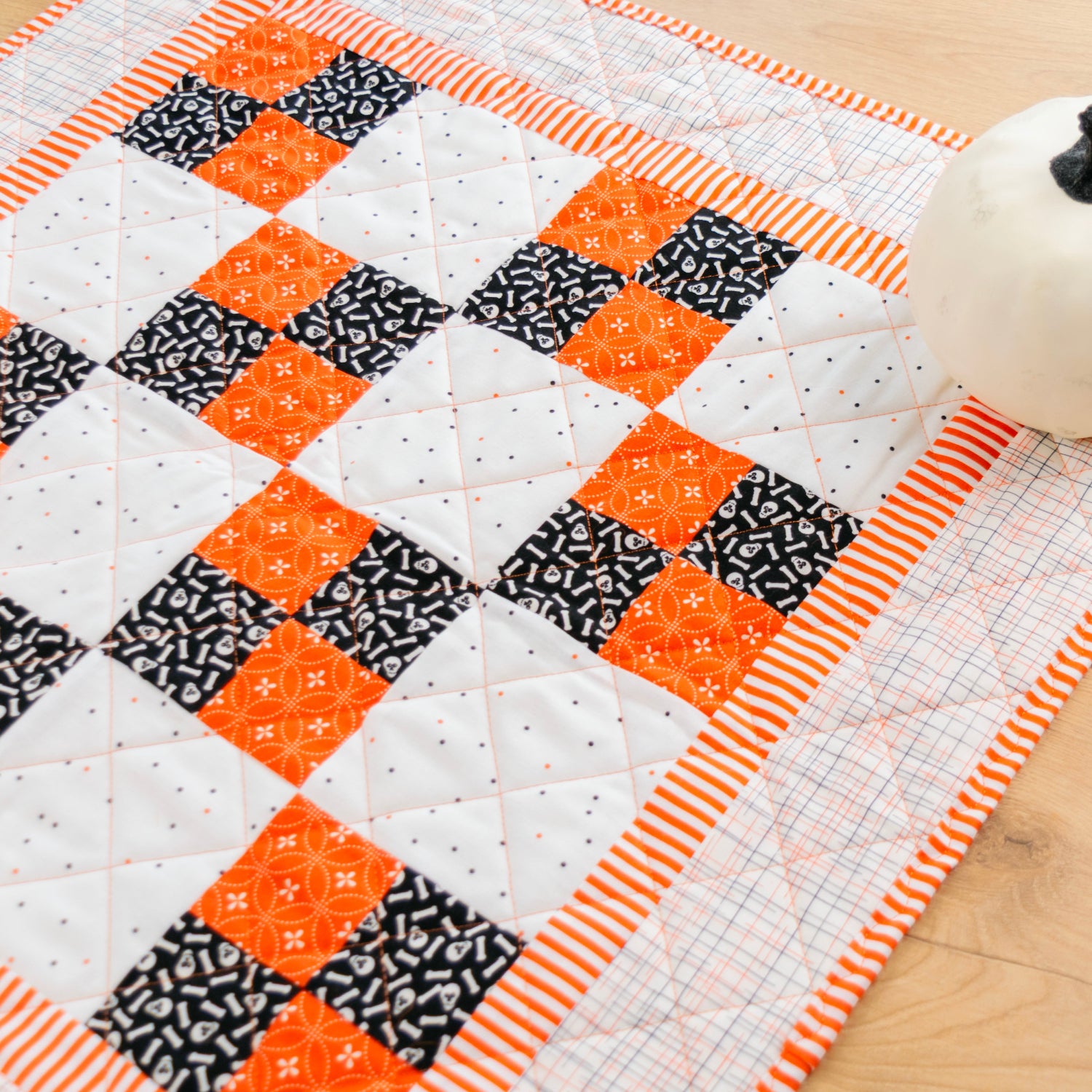 A handmade quilted table runner or placemat featuring a geometric patchwork design of orange, black, and white fabrics, styled on a light wood surface next to a small white decorative pumpkin.