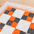 A close-up of a handmade quilted table runner or mat featuring a patchwork design of orange, black, and white fabrics, laid out on a light wood surface.