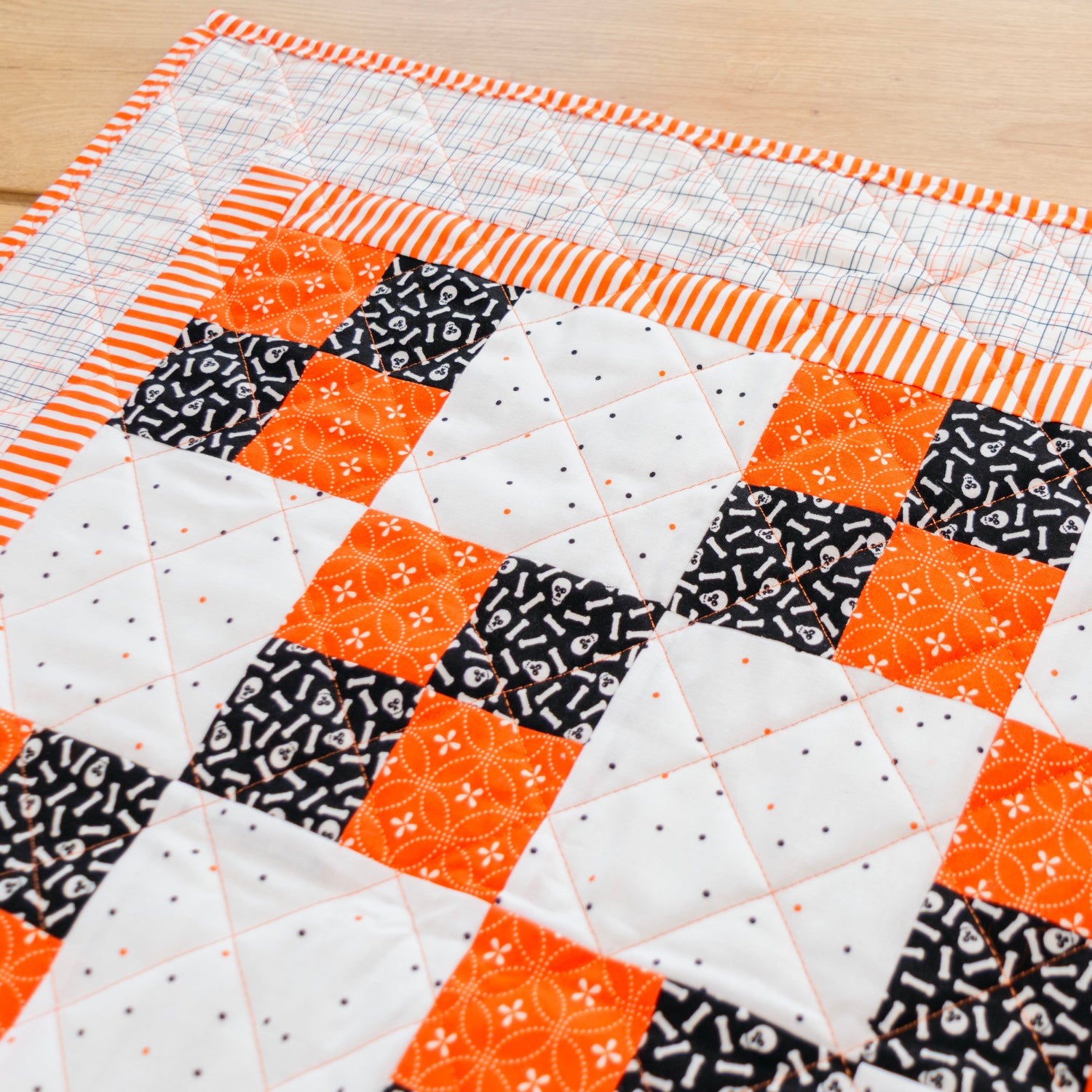 A close-up of a handmade quilted table runner or mat featuring a patchwork design of orange, black, and white fabrics, laid out on a light wood surface.