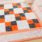 A handmade quilted table runner or placemat featuring a geometric patchwork design of orange, black, and white blocks, bordered by orange and white striped fabric, laid out on a light wood surface.