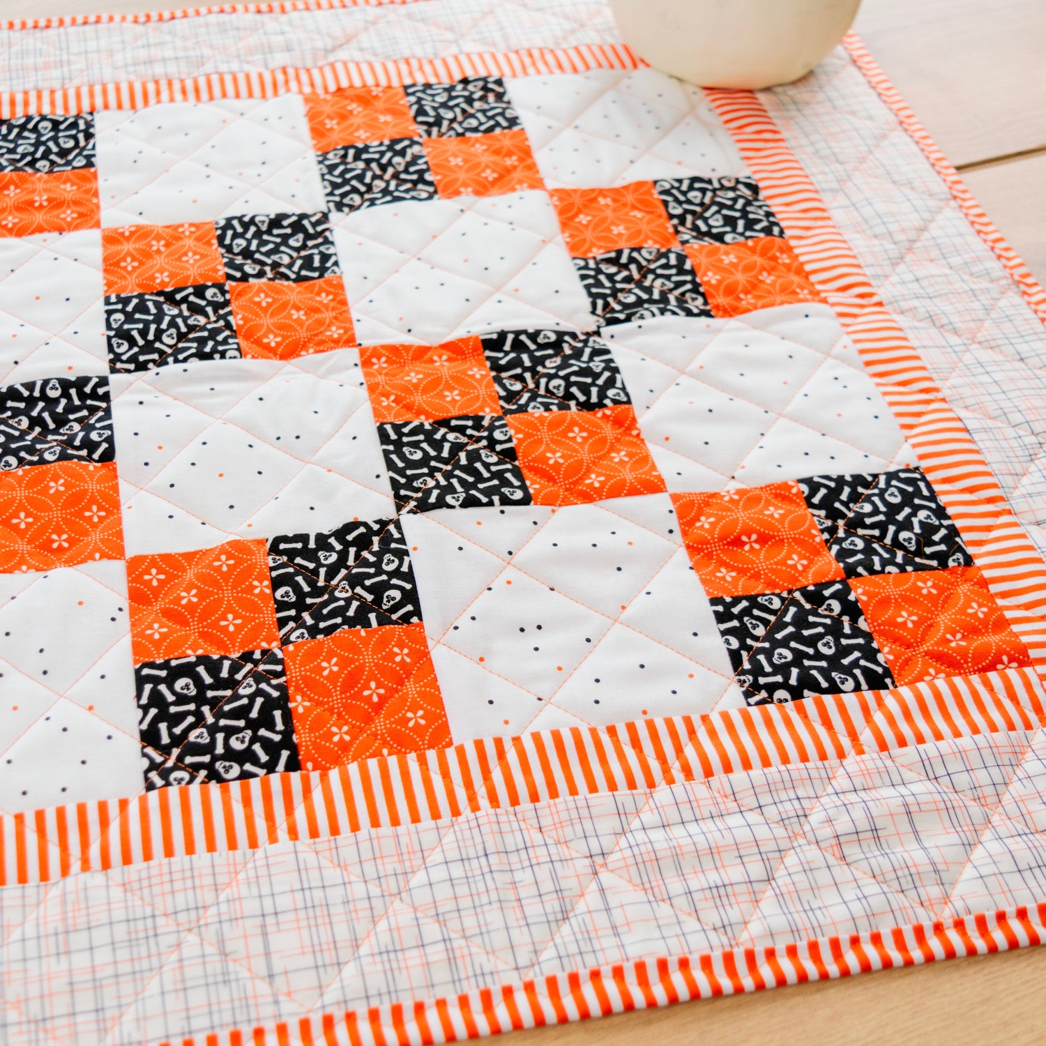 A handmade quilted table runner or placemat featuring a geometric patchwork design of orange, black, and white blocks, bordered by orange and white striped fabric, laid out on a light wood surface.