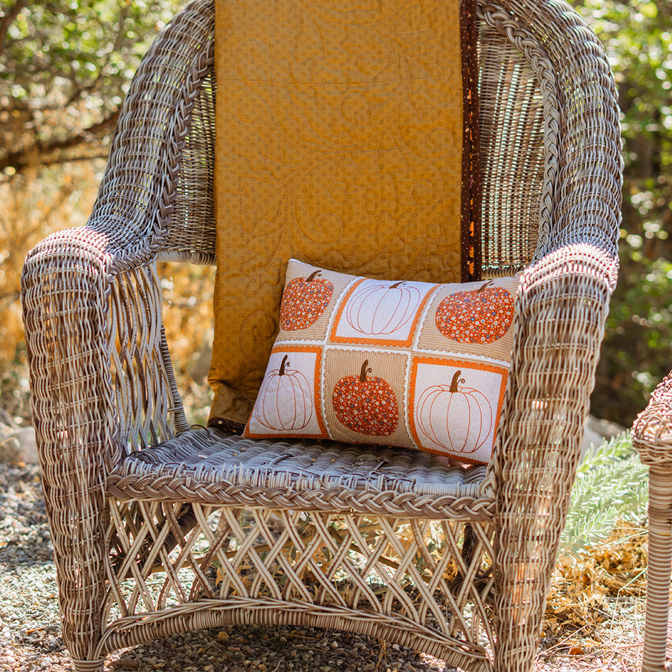 A square decorative throw pillow with a pumpkin pattern, styled on the seat of a wicker outdoor chair with a yellow quilted blanket in a garden setting.