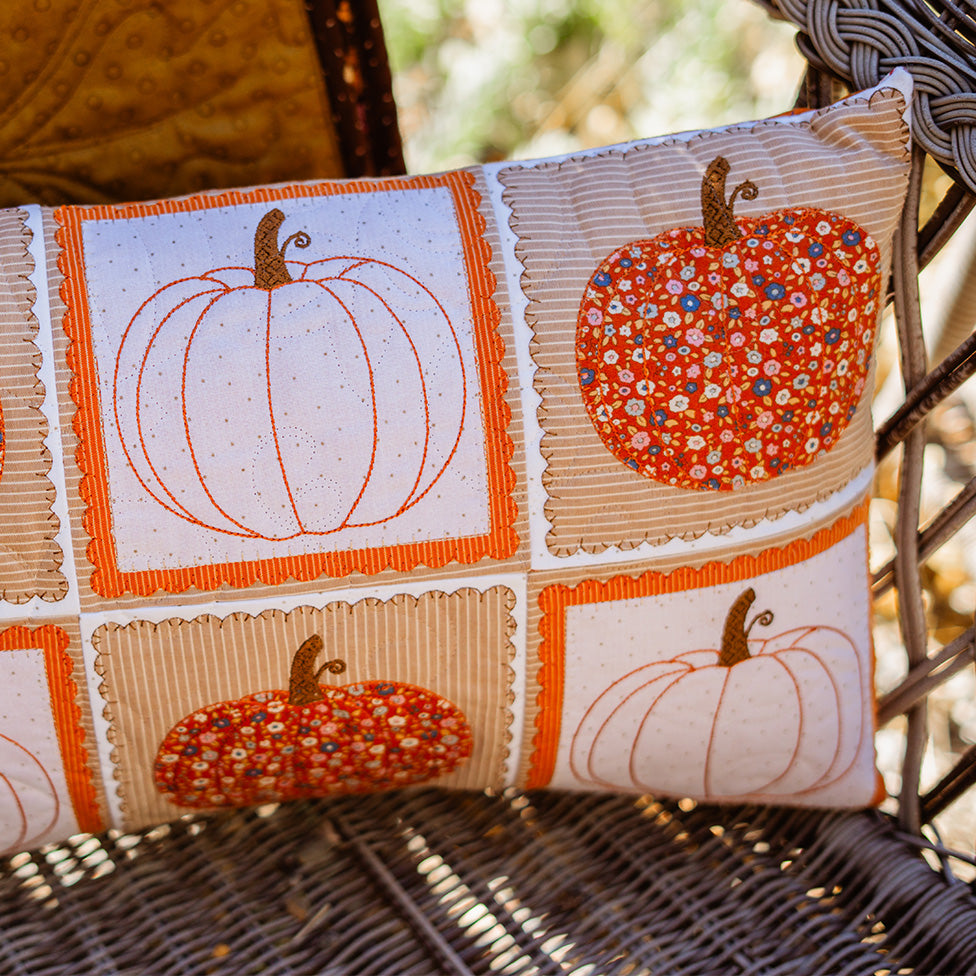 A square decorative throw pillow with an autumn patchwork design featuring embroidered and floral pumpkin motifs in orange, beige, and white, resting on a wicker chair in an outdoor setting.