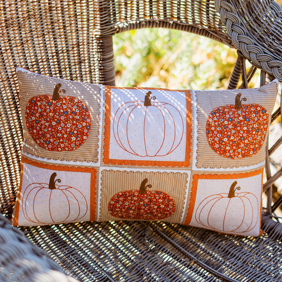 A rectangular lumbar throw pillow with a 3x2 grid of autumn pumpkin designs, resting on a woven wicker chair. The pillow features a cream background with orange borders and six pumpkins in solid orange, white, and orange-and-white floral patterns.