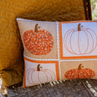 A rectangular decorative throw pillow with a patchwork autumn pumpkin design, resting on a wicker chair next to a yellow quilted cushion.