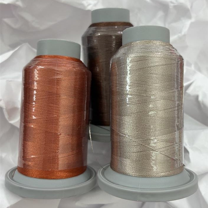 Three spools of thread on gray plastic cones, arranged together on crumpled white paper. The front-left spool contains copper-orange thread, the front-right spool contains champagne-beige thread, and the back spool contains dark brown thread.