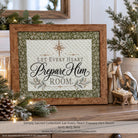 A framed decorative sign reading “Let Every Heart Prepare Him Room” displayed on a shelf with holiday greenery, candles, and a small nativity figurine. The sign has a quilted cream center, a green patterned border, and a rustic brown wood frame.