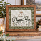 A framed quilted sign with the text 'Let Every Heart Prepare Him Room' in decorative lettering, surrounded by a green floral border and a medium brown wooden frame. The sign is displayed on a wooden shelf next to a lit candle, books, and a potted plant.