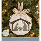 A round embroidered Christmas ornament hanging on a decorated Christmas tree. The ornament features a nativity scene with Mary, Joseph, and baby Jesus inside a stable design, tied with a beige and gold ribbon.