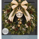 A festive Christmas wreath hanging on a dark gray door, decorated with green pine branches, white berries, brown pinecones, gold bells, and a large cream and olive bow with a nativity ornament. Text at the bottom identifies it as a Nativity Christmas Ornament from the Simply Sacred Collection.