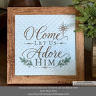 A framed decorative sign with the phrase “O Come Let Us Adore Him” in brown script and block lettering on a light blue quilted background. The sign is housed in a rustic brown wooden frame, styled with evergreen branches on a dark wooden surface.