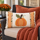 A rectangular decorative throw pillow with a pumpkin design and pom-pom trim, styled on a wicker patio chair with an orange knit blanket and fall decor in a porch setting.