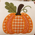 A close-up of a decorative fabric pillow featuring an applique pumpkin design. The pumpkin has orange and cream gingham patterned sections, orange embroidered borders, a dark brown stem, and a green leaf with embroidered details.