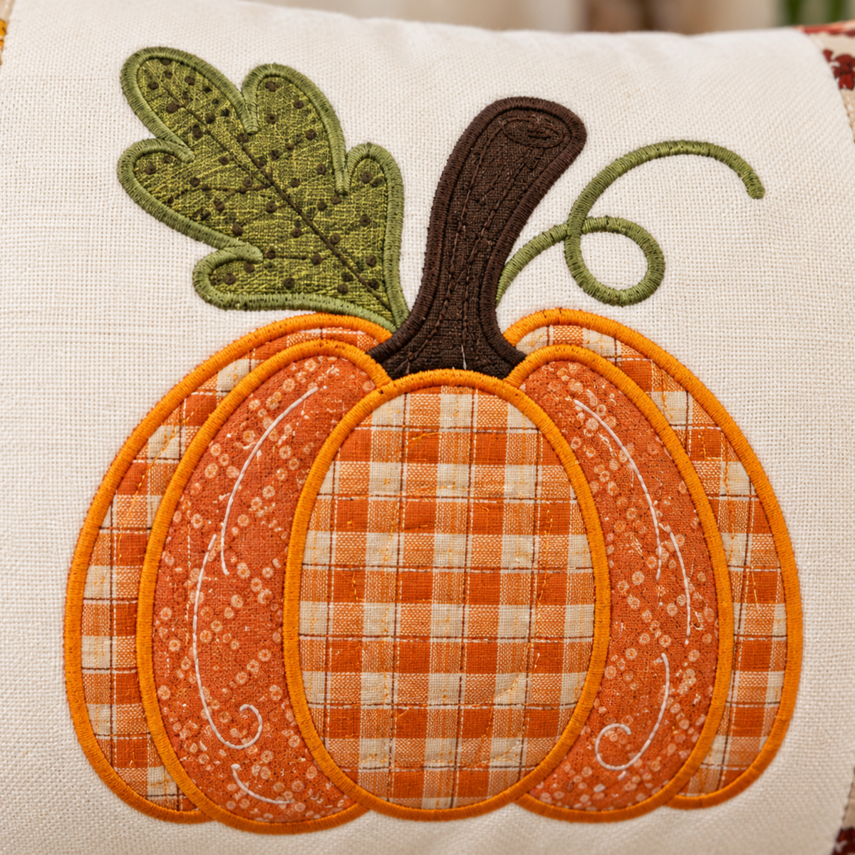 A close-up of a decorative fabric pillow featuring an applique pumpkin design. The pumpkin has orange and cream gingham patterned sections, orange embroidered borders, a dark brown stem, and a green leaf with embroidered details.
