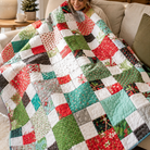 A woman sitting on a beige sofa wrapped in a large patchwork quilted blanket with red, green, white, and gray patterned squares, in a cozy living room setting.
