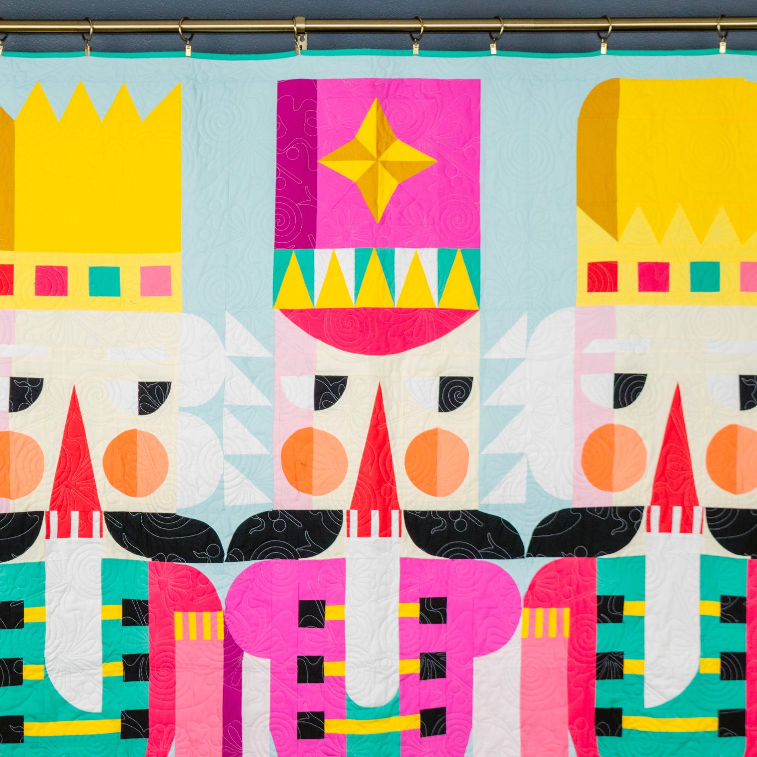 A colorful quilted wall hanging or tapestry featuring three stylized nutcracker figures in bright geometric patterns, suspended from a metal rod against a dark gray wall.