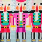 A colorful quilted wall hanging or blanket featuring three stylized nutcracker figures in bright pink, red, teal, black, white, and yellow on a pale blue background, shown hanging indoors against a dark gray wall with a small tree and red ladder visible at the edges.