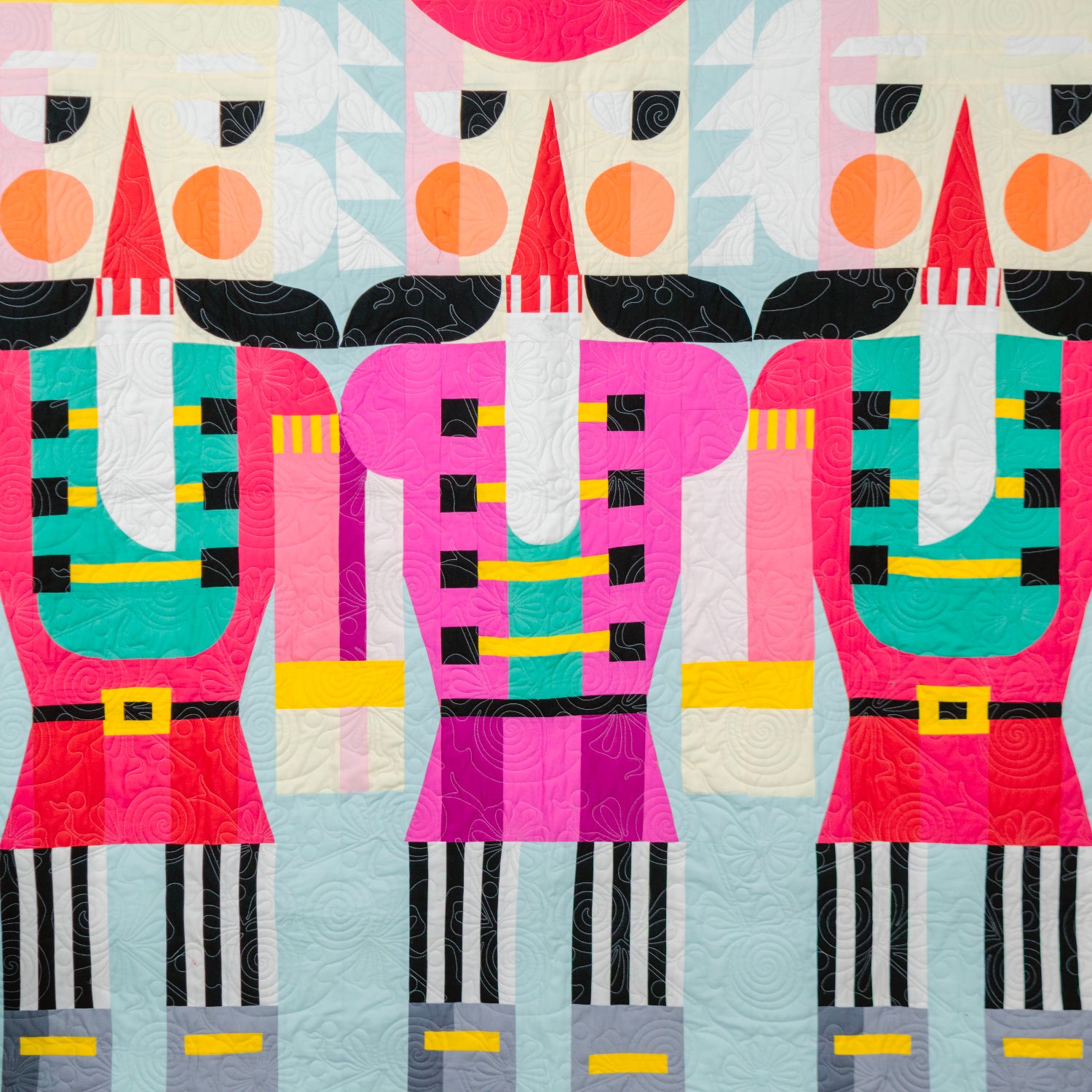 A colorful quilted wall hanging or blanket featuring three stylized nutcracker figures in bright pink, red, teal, black, white, and yellow on a pale blue background, shown hanging indoors against a dark gray wall with a small tree and red ladder visible at the edges.