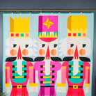 A colorful quilt featuring three stylized nutcracker figures with bright pink, yellow, teal, black, and white geometric patterns on a light blue background, hanging on a rod against a dark gray wall.