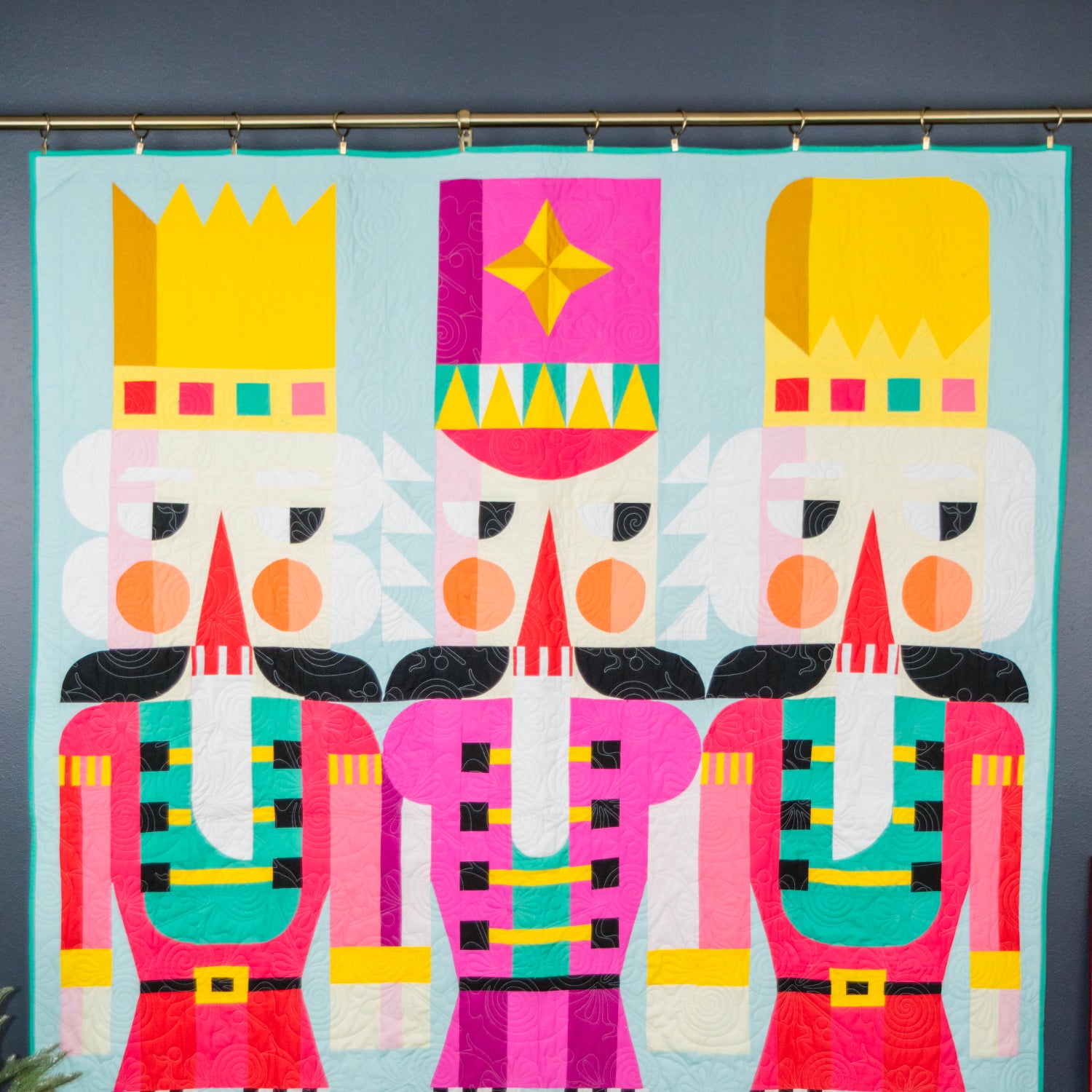 A colorful quilt featuring three stylized nutcracker figures with bright pink, yellow, teal, black, and white geometric patterns on a light blue background, hanging on a rod against a dark gray wall.