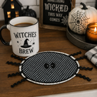 A Halloween-themed tabletop scene with a white ceramic mug printed with the words “WITCHES BREW” and a black witch hat graphic, placed beside a round black-and-white gingham spider placemat on a wooden table. The setting includes a lit candle, a white pumpkin with a spiderweb print, and a sign reading “Something Wicked This Way Comes.