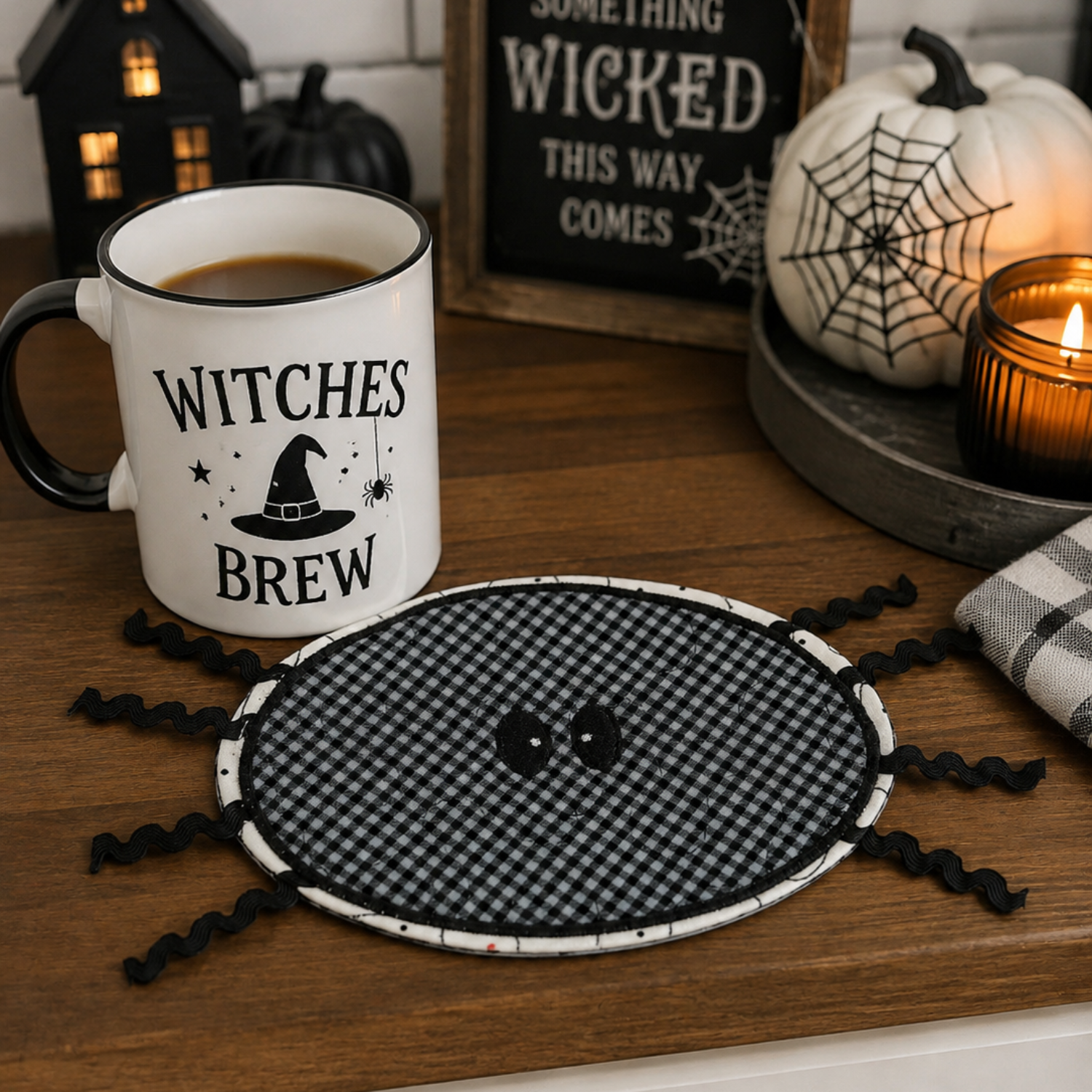 A Halloween-themed tabletop scene with a white ceramic mug printed with the words “WITCHES BREW” and a black witch hat graphic, placed beside a round black-and-white gingham spider placemat on a wooden table. The setting includes a lit candle, a white pumpkin with a spiderweb print, and a sign reading “Something Wicked This Way Comes.
