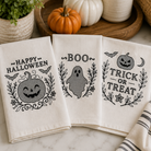 Three white kitchen towels with black Halloween-themed prints are arranged on a marble countertop. The towels feature 'Happy Halloween', 'Boo', and 'Trick or Treat' designs with pumpkins, ghosts, bats, and leaves.