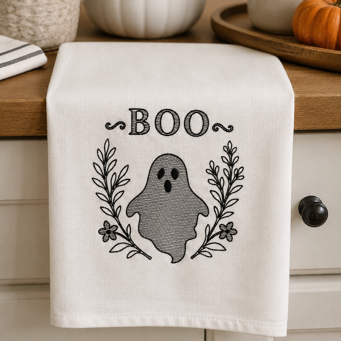An off-white kitchen towel with a black embroidered 'BOO' design featuring a ghost and leafy wreath, hanging on a wooden kitchen counter with seasonal props like pumpkins and a plant.